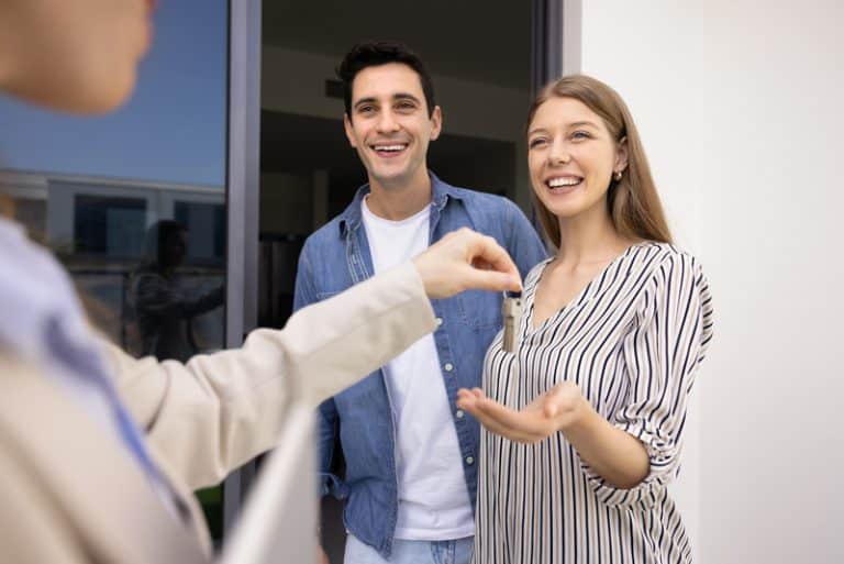 Happy couple buying new house, getting key from realtor, meeting with seller, real estate agent outdoors at apartment entrance. Positive young man and woman renting flat